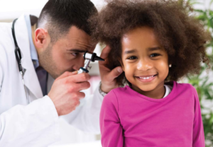 A provider using an otoscope examines the ear of a smiling little girl wearing a pink shirt